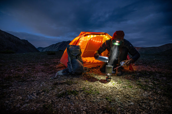 A camper prepares food with a headlamp near an orange tent lit with string lights at dusk, surrounded by mountains and rocky terrain. A backpack sits nearby.