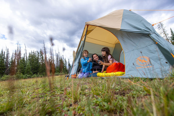 Three children sit inside a tent on a grassy field, smiling and looking at a phone together, with trees and a cloudy sky in the background.