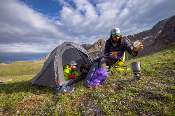 Two people camp on a grassy mountain slope. One sits in a tent, the other cooks with a camping stove beside the tent. Mountains and a partly cloudy sky are in the background.