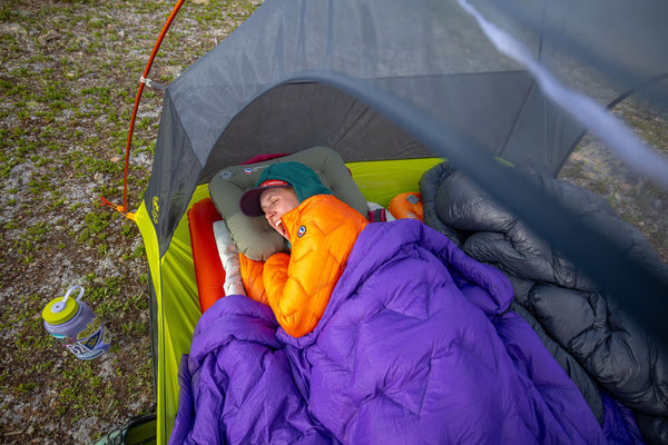 A person wearing a green hat and orange jacket sleeps in a tent, wrapped in a purple sleeping bag with camping gear nearby and a water bottle outside the tent on the grass.