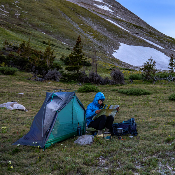 A person in a blue jacket sits beside the spacious Big Agnes Europe Pitchpine VST 1.5 ultralight tent on grassy mountains, reading a map, with camping gear nearby and snow patches visible on the hillside in the background.