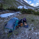Two people and a small dog relax beside the Big Agnes Europe Sarvis VST 3 ultralight shelter in a snowy, mountainous area with trees. Camping gear is scattered on the grassy, rocky ground around them.