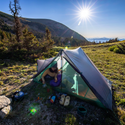A camper relaxes inside a Big Agnes Europe String Ridge VST 2.5 tent on a grassy hillside at sunrise, camping gear nearby and morning light illuminating distant mountains and trees.