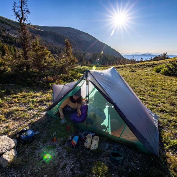 A camper relaxes inside a Big Agnes Europe String Ridge VST 2.5 tent on a grassy hillside at sunrise, camping gear nearby and morning light illuminating distant mountains and trees.