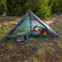A person relaxes on a sleeping pad inside the Big Agnes Europe String Ridge VST 1.5 ultralight tent, reading a device. Shoes, water bottle, and camping gear rest outside the tent on grassy ground among trees and greenery.