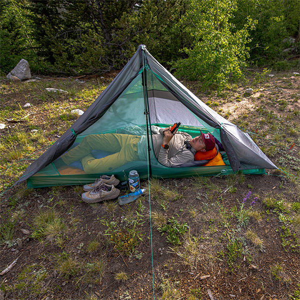 A person relaxes on a sleeping pad inside the Big Agnes Europe String Ridge VST 1.5 ultralight tent, reading a device. Shoes, water bottle, and camping gear rest outside the tent on grassy ground among trees and greenery.